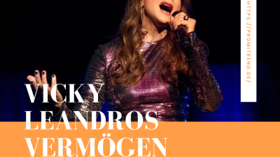 Person mit langen Haaren in glitzerndem Outfit hält ein Mikrofon auf der Bühne. Text "VICKY LEANDROS VERMÖGEN" auf orangefarbenem Hintergrund. Vertikaler Text "HTTPS://PROMITREND.DE/".