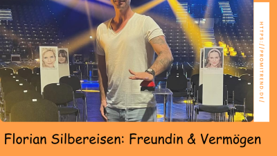 Ein Mann in einem weißen T-Shirt steht in einer leeren Veranstaltungshalle mit Scheinwerfern im Hintergrund. Der Text "Florian Silbereisen: Freundin & Vermögen" ist unten auf dem Bild zu sehen.