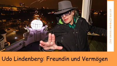 Eine Person mit verdecktem Gesicht steht auf einem Balkon bei Nacht, im Hintergrund sind Feuerwerkskörper und eine beleuchtete Kugel zu sehen. Der Text auf dem Bild lautet: "Udo Lindenberg: Freundin und Vermögen".